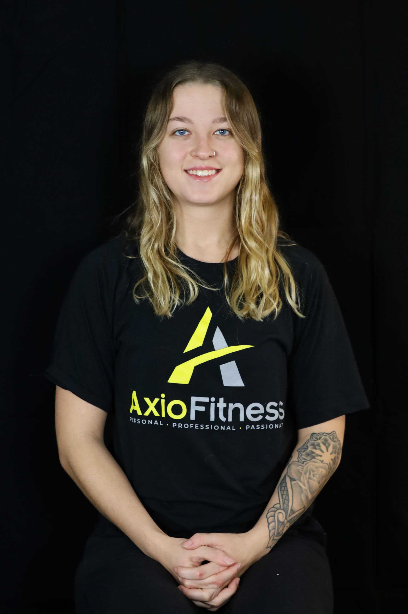 Woman with blonde hair and a tattoo, wearing a black AxioFitness t-shirt, smiles at the camera against a black backdrop.