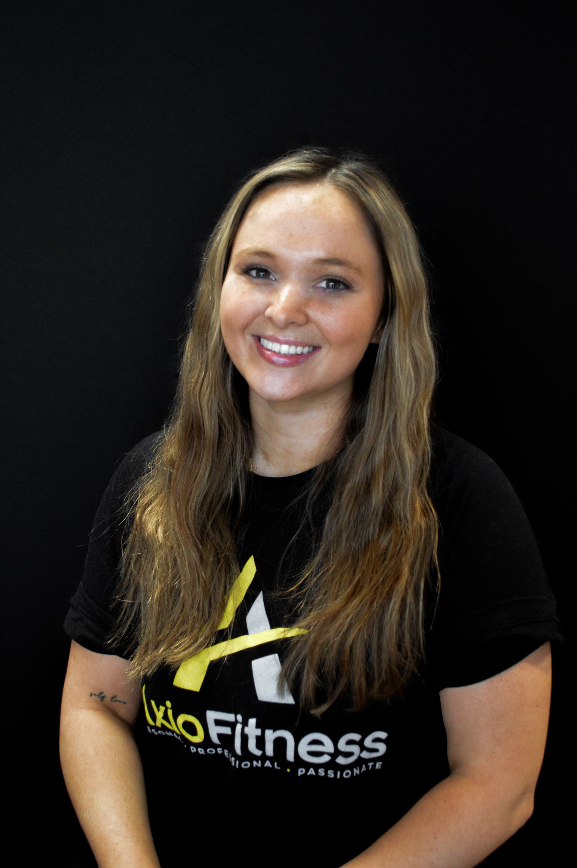 Woman with long blonde hair, wearing a black t-shirt, smiling at the camera. Black background.