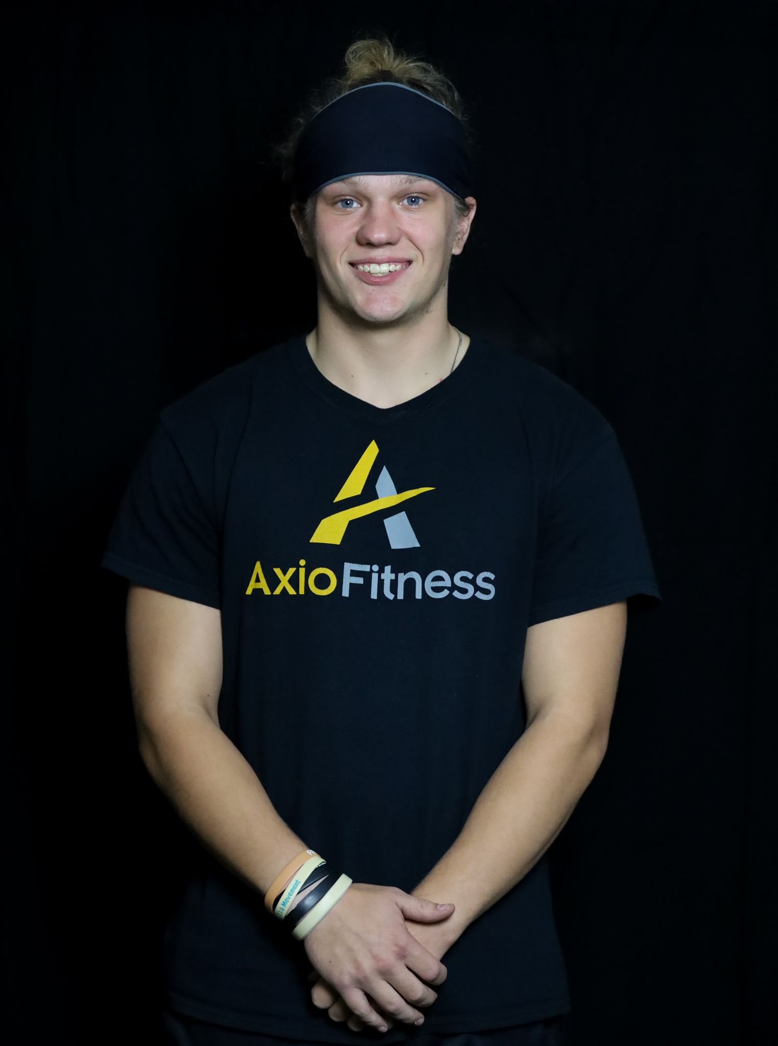 Young man wearing a black AxioFitness t-shirt and headband smiles, arms crossed, against a black backdrop.
