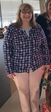 A smiling woman with glasses wearing a patterned button-down shirt, light pink pants, and black shoes.