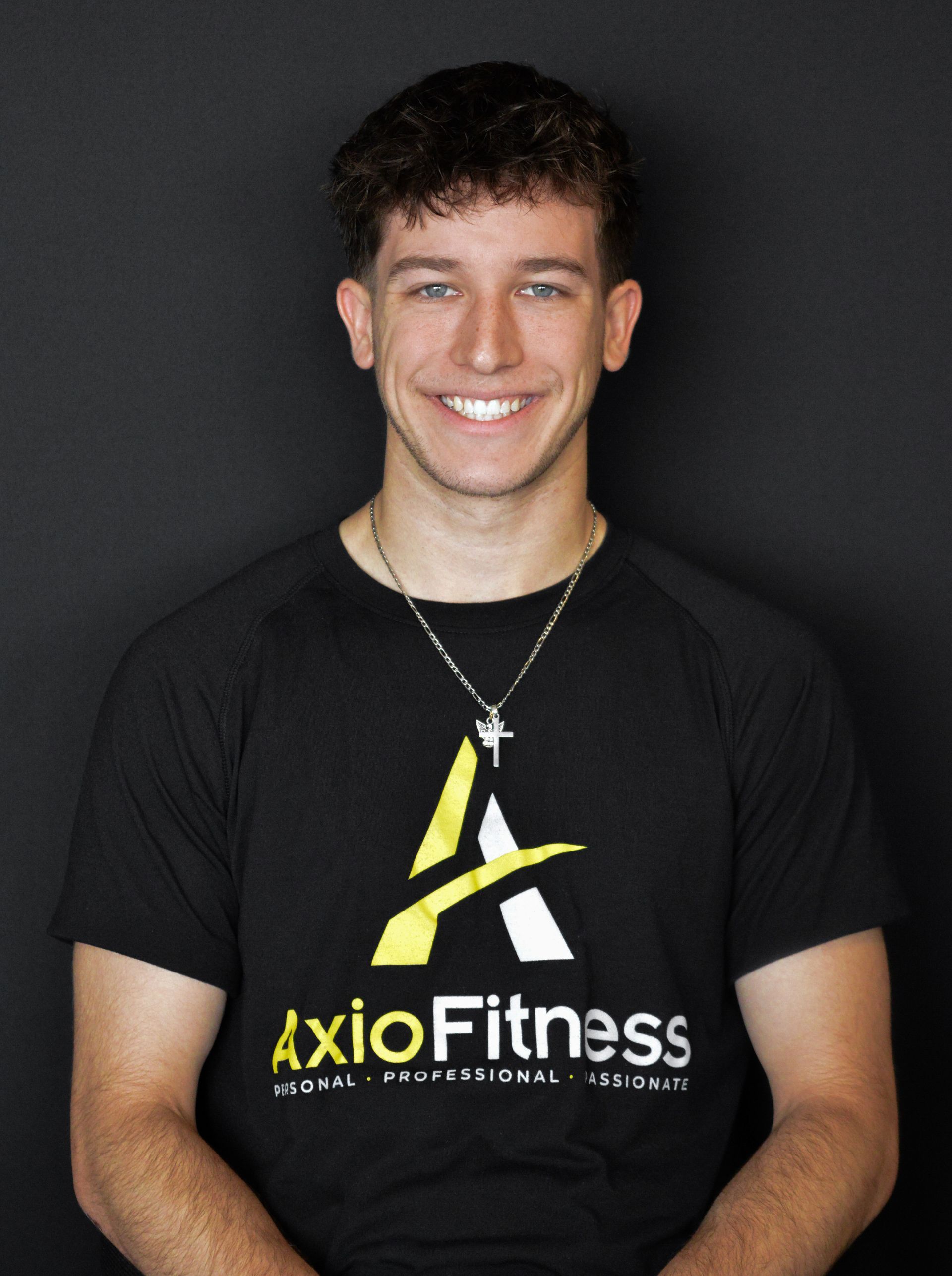 Smiling young man wearing a black AxioFitness t-shirt and a silver cross necklace against a black background.