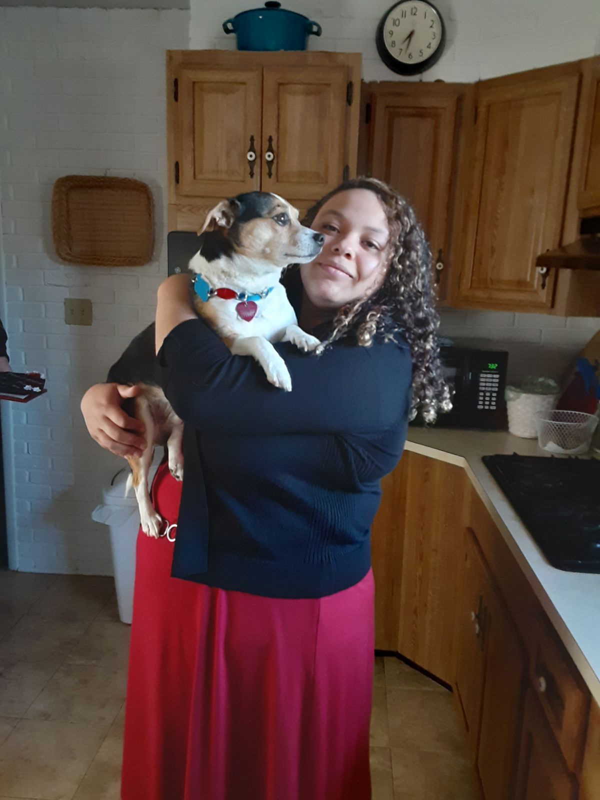 Woman in red skirt holds a small dog in a kitchen, smiling. The dog is wearing a collar.