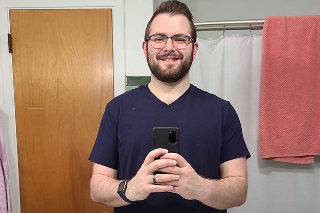 Man with glasses and a beard smiles while holding a phone in a bathroom. He wears a dark blue v-neck shirt.