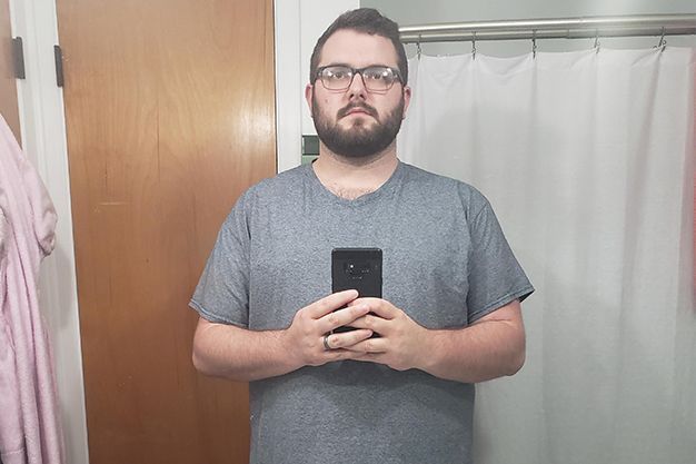 Man with glasses and a beard taking a mirror selfie in a bathroom, holding a phone. He wears a gray t-shirt.