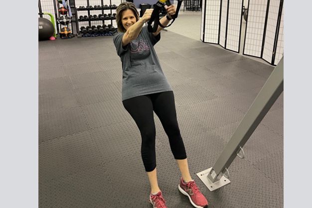 Woman in gym using TRX suspension trainer, holding handles and leaning back.