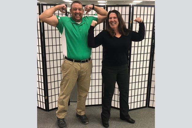 A man and a woman flexing their biceps. The man wears a green shirt and khaki pants, while the woman wears a black shirt.
