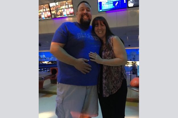 A smiling couple poses together at a bowling alley. The man, wearing a blue shirt, has his hand on his stomach.