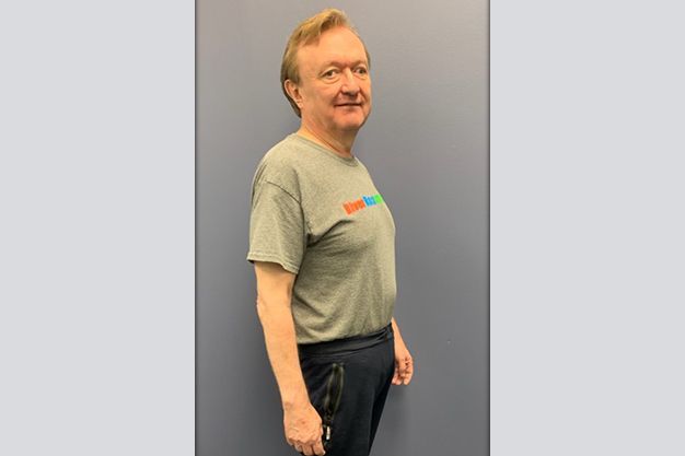 A man with light hair in a grey t-shirt and dark pants stands facing left. He smiles in front of a blue wall.
