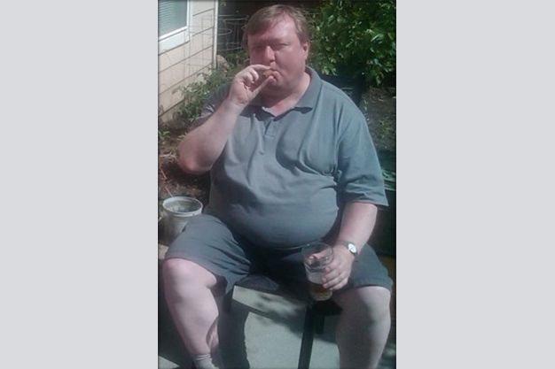 Overweight man sitting outside, smoking a cigarette and holding a glass. He wears a grey polo shirt and shorts.