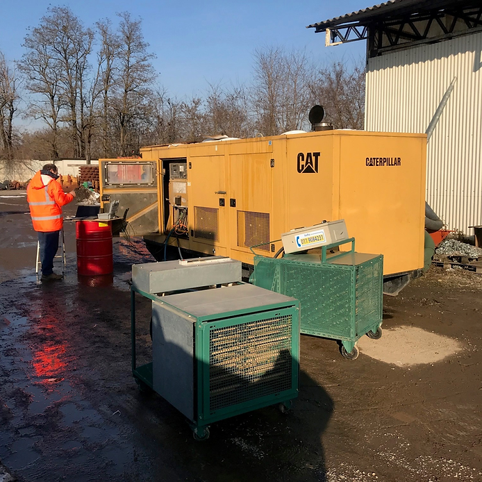 Un operaio con una giacca arancione vicino a un generatore CAT giallo, un carrello verde e un barile all'aperto.
