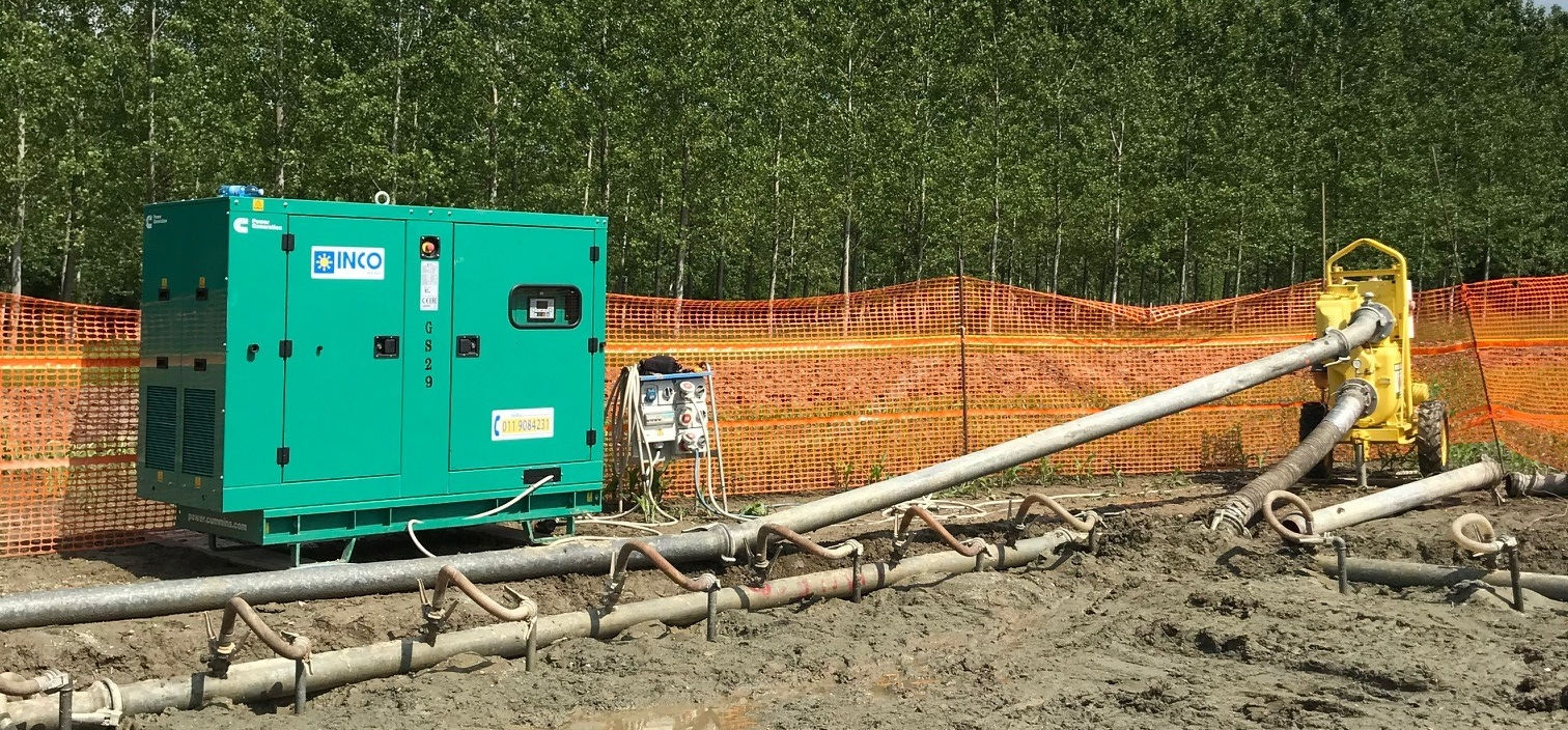 Un generatore verde e tubi in un cantiere con recinzione di sicurezza arancione e alberi sullo sfondo.
