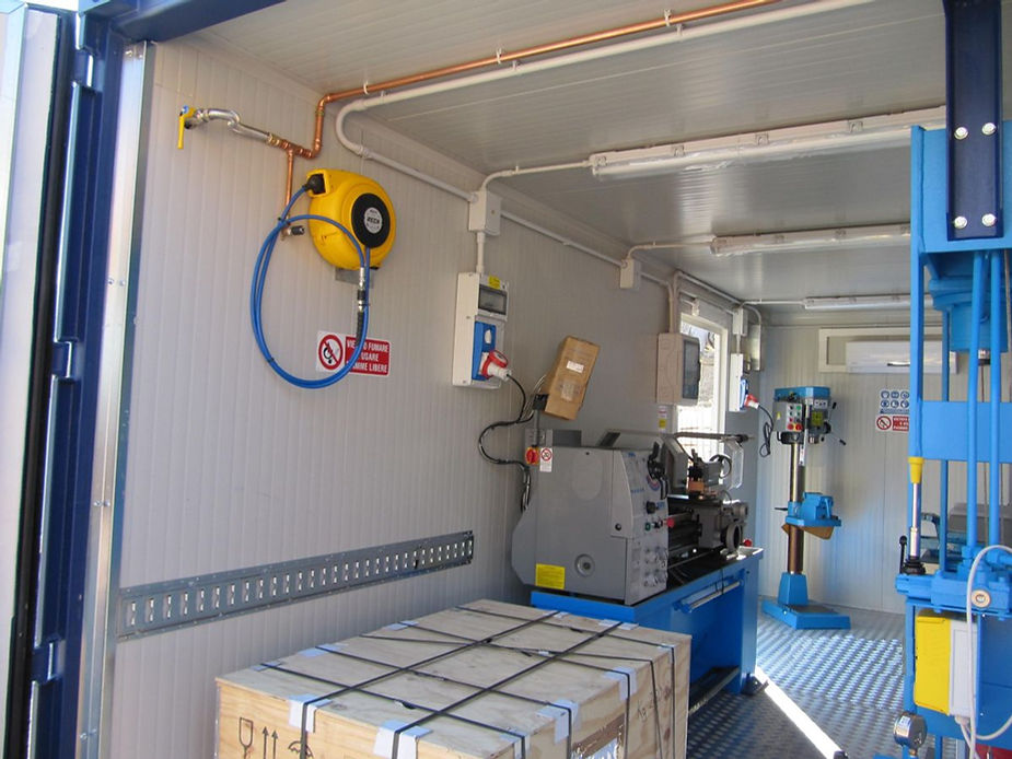 Interno di un container da officina con utensili: tornio, trapano a colonna, tubo dell'aria e scatole.