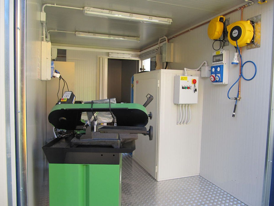 Interno di un'officina container con una sega a nastro verde, un quadro elettrico e un tubo dell'aria retrattile.