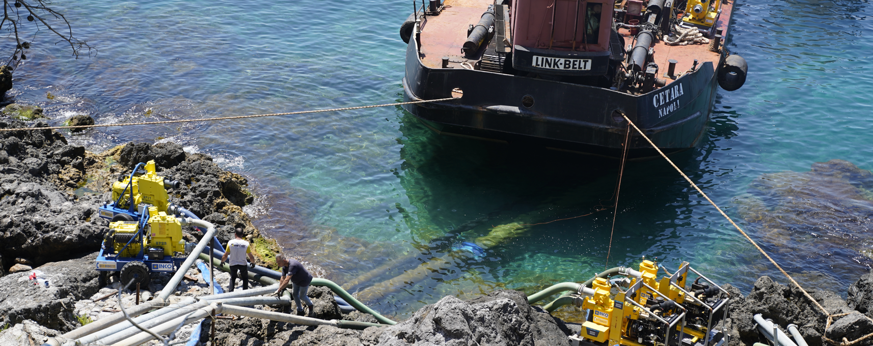 Imbarcazione attraccata vicino alla costa rocciosa con operai e attrezzature sul posto.