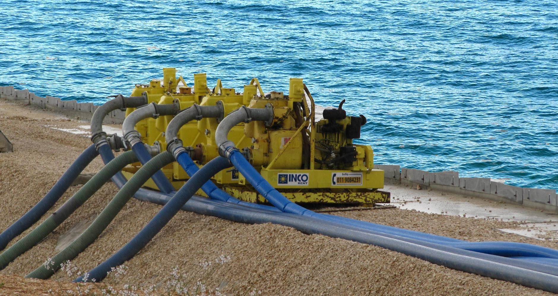 Pompe industriali gialle con tubi flessibili su una costa rocciosa, acqua sullo sfondo.
