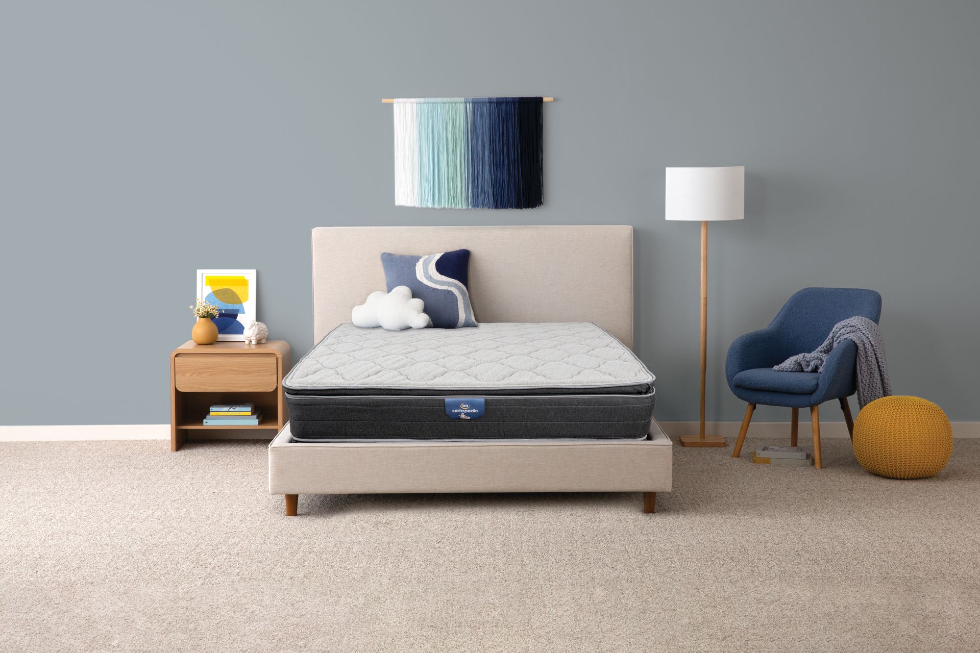 Dormitorio con cama, mesita de noche, sillón y decoración. Cuenta con un colchón sobre una base de cama beige, una pared azul y una alfombra neutra.