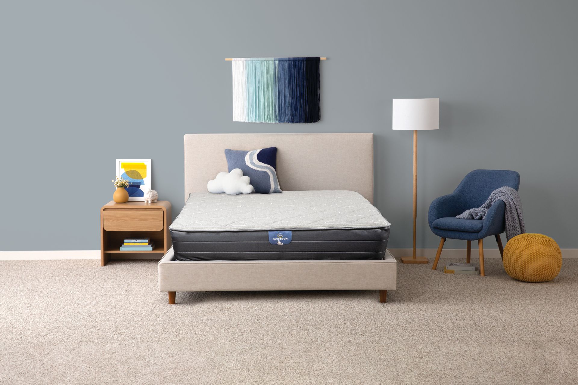Escena de dormitorio: cama con colchón gris claro, silla azul, lámpara de pie, mesita de noche, arte de pared, alfombra neutra y pared.