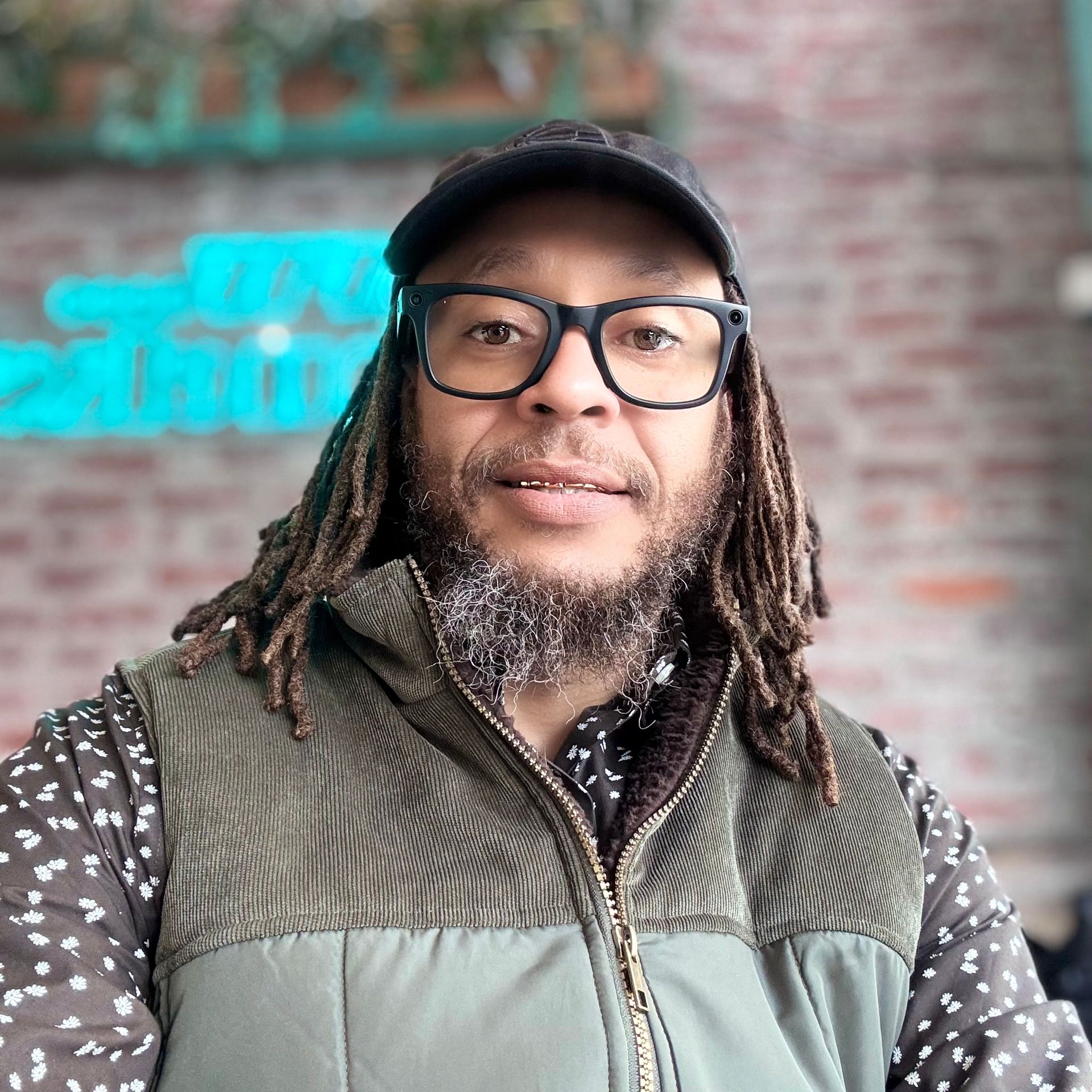 Person wearing glasses and a cap, taking a selfie against a brick wall with turquoise signage.