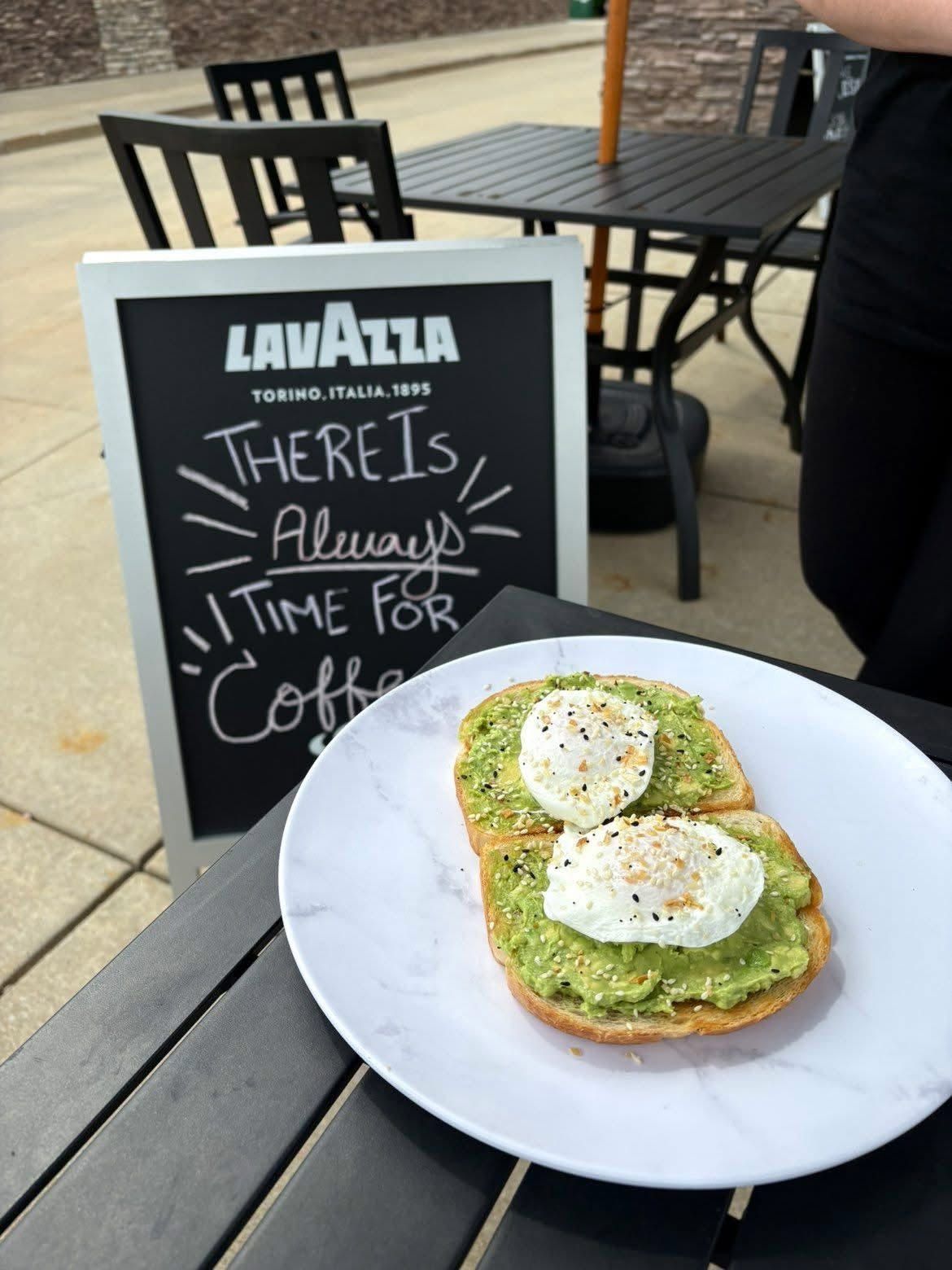 Avocado toast with poached eggs on a white plate beside a café chalkboard sign about coffee.