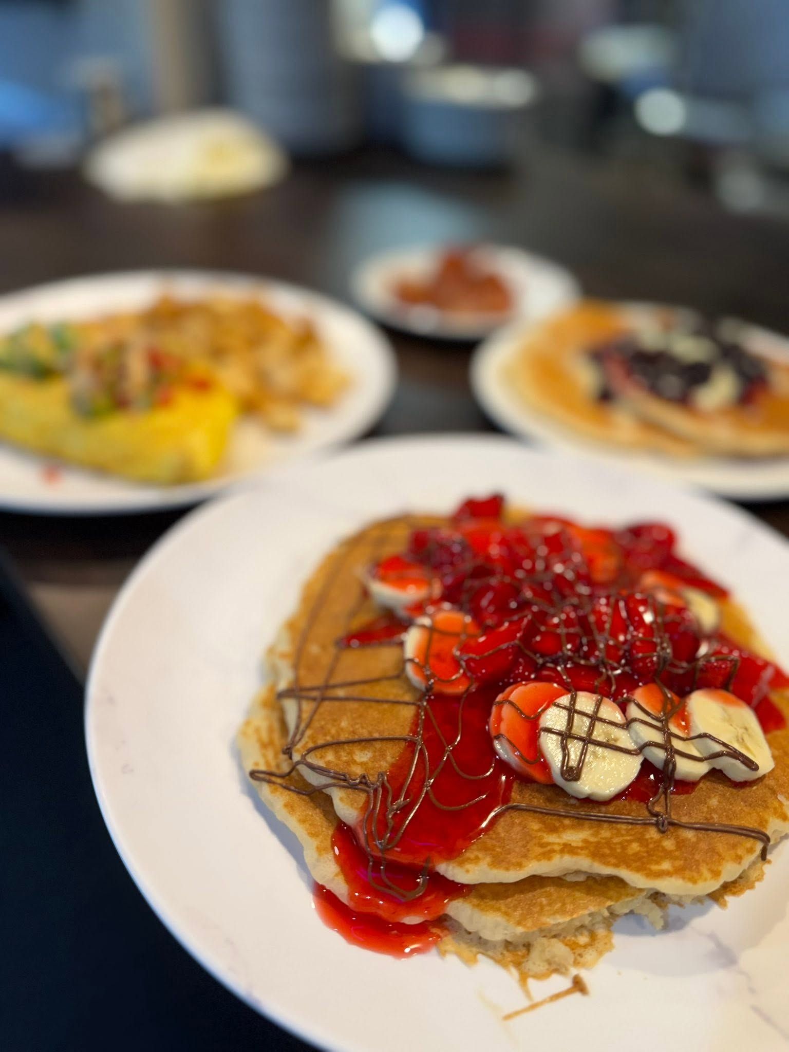 Stack of pancakes topped with strawberries and syrup on a white plate, with blurred breakfast dishes behind