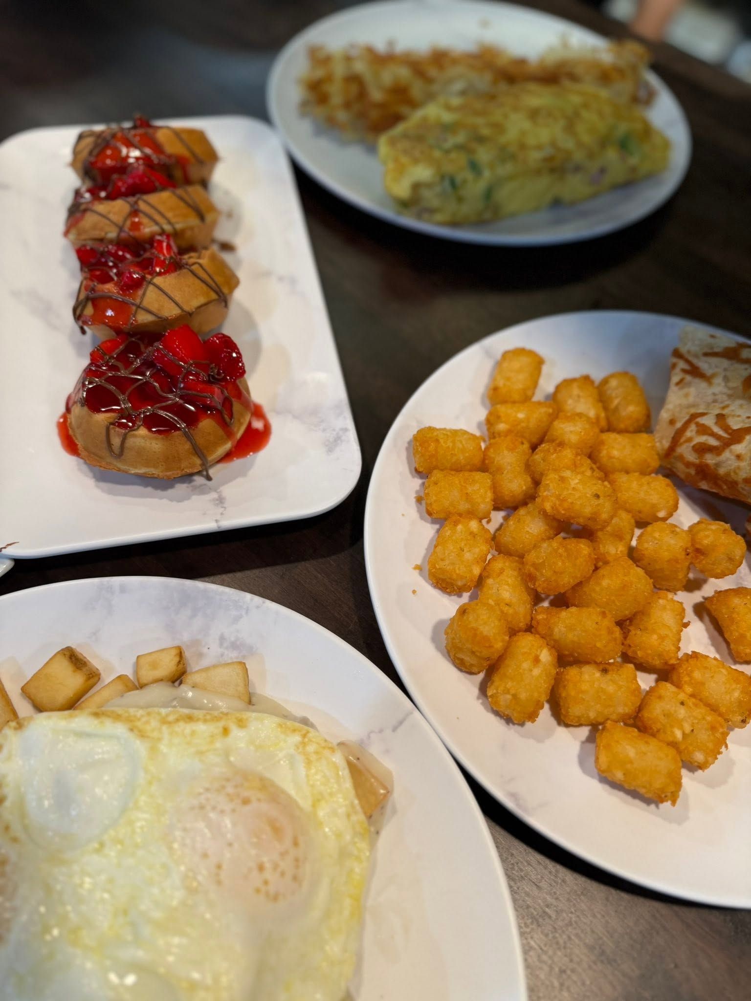 Assorted breakfast dishes on white plates, including fried eggs, tater tots, toast, and a fruit-topped pastry.