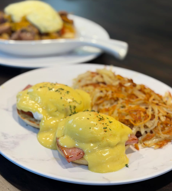Eggs Benedict with hollandaise sauce and hash browns on a white plate, with a second plate blurred behind.