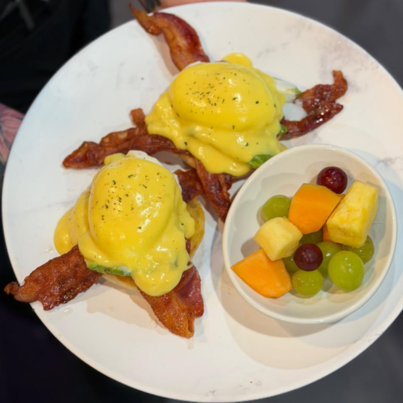 Bacon eggs Benedict on a white plate with a small fruit cup of grapes, pineapple, and melon