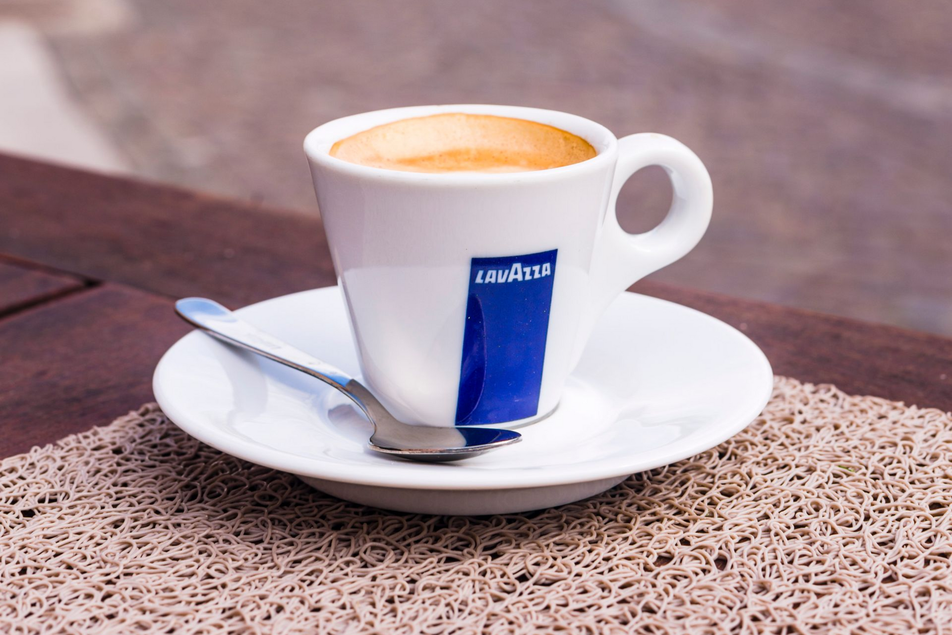 White espresso cup on saucer with spoon and blue logo, filled with coffee on a stone table.