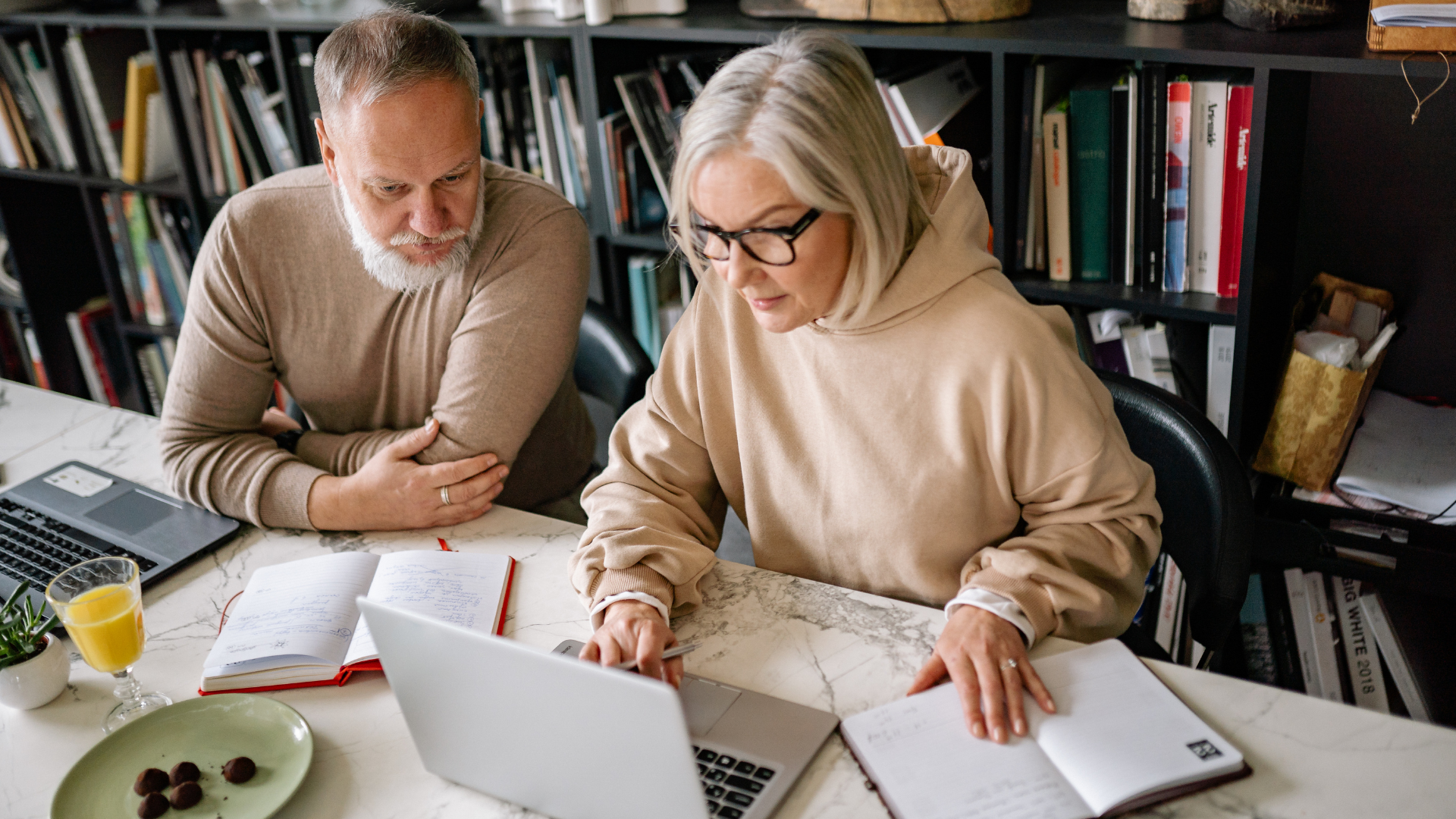 Two people sitting at a table with laptops and journals, collaborating in a home office with bookshelves in the background.
