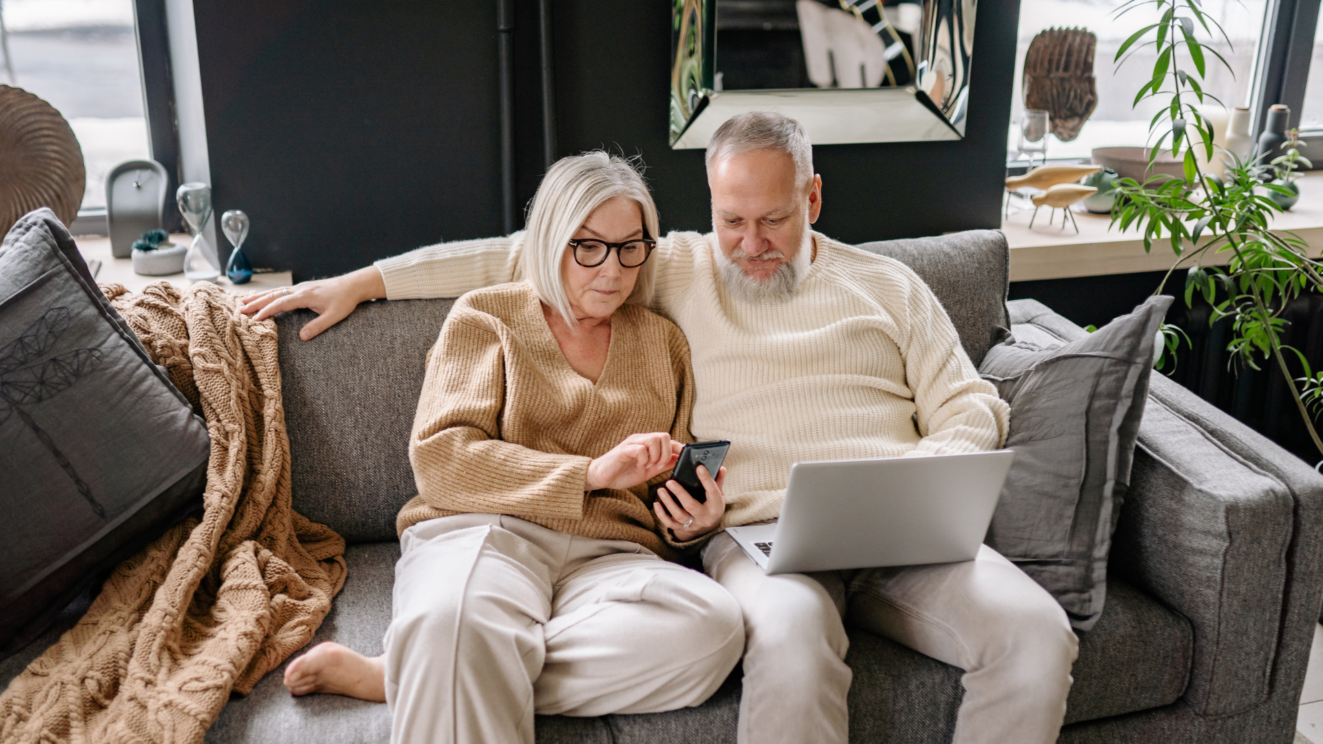 Two people sit together on a gray sofa, looking at a laptop and smartphone in a modern, plant-filled living room.