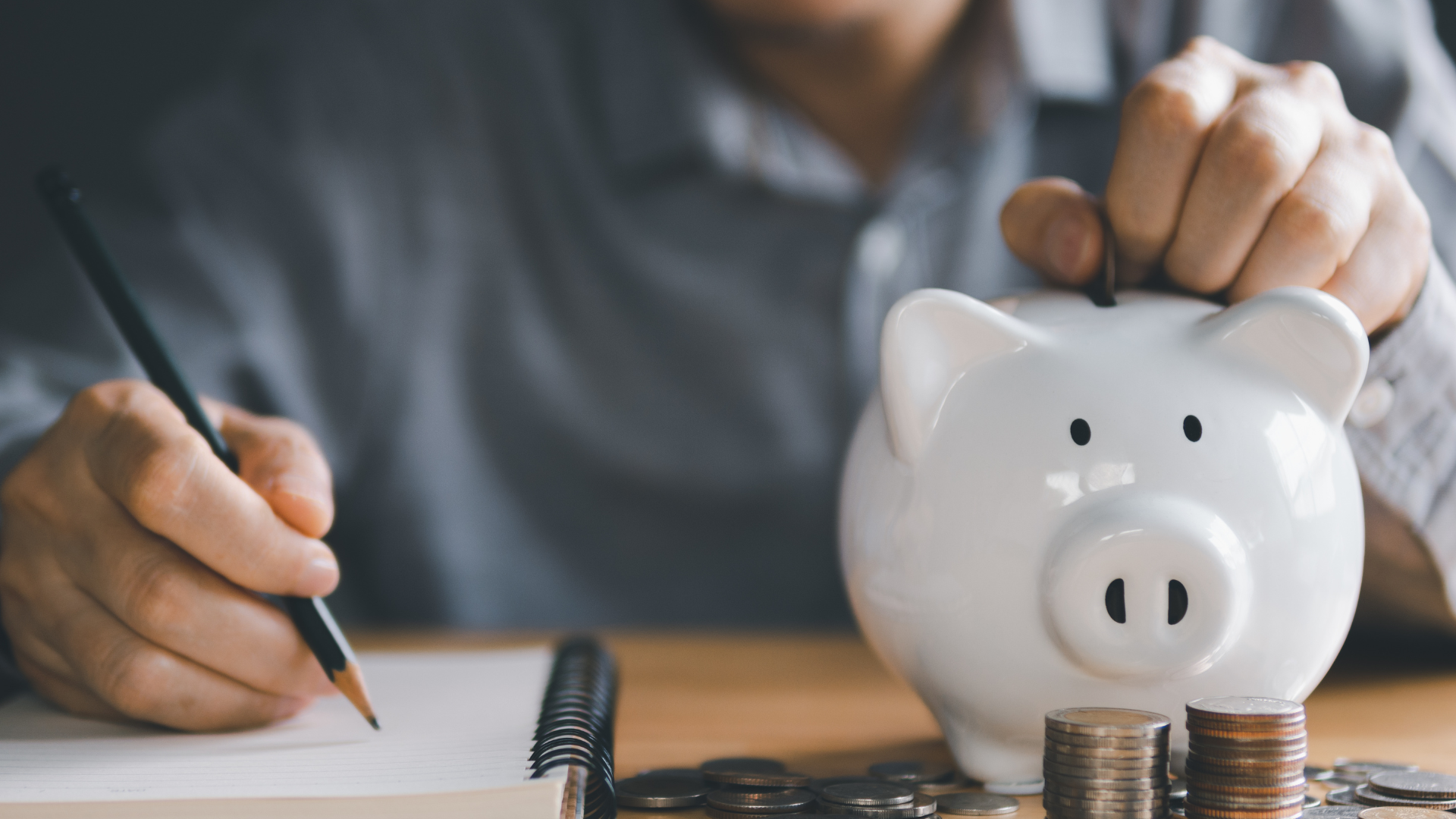 A man is putting coins into a piggy bank while writing in a notebook.