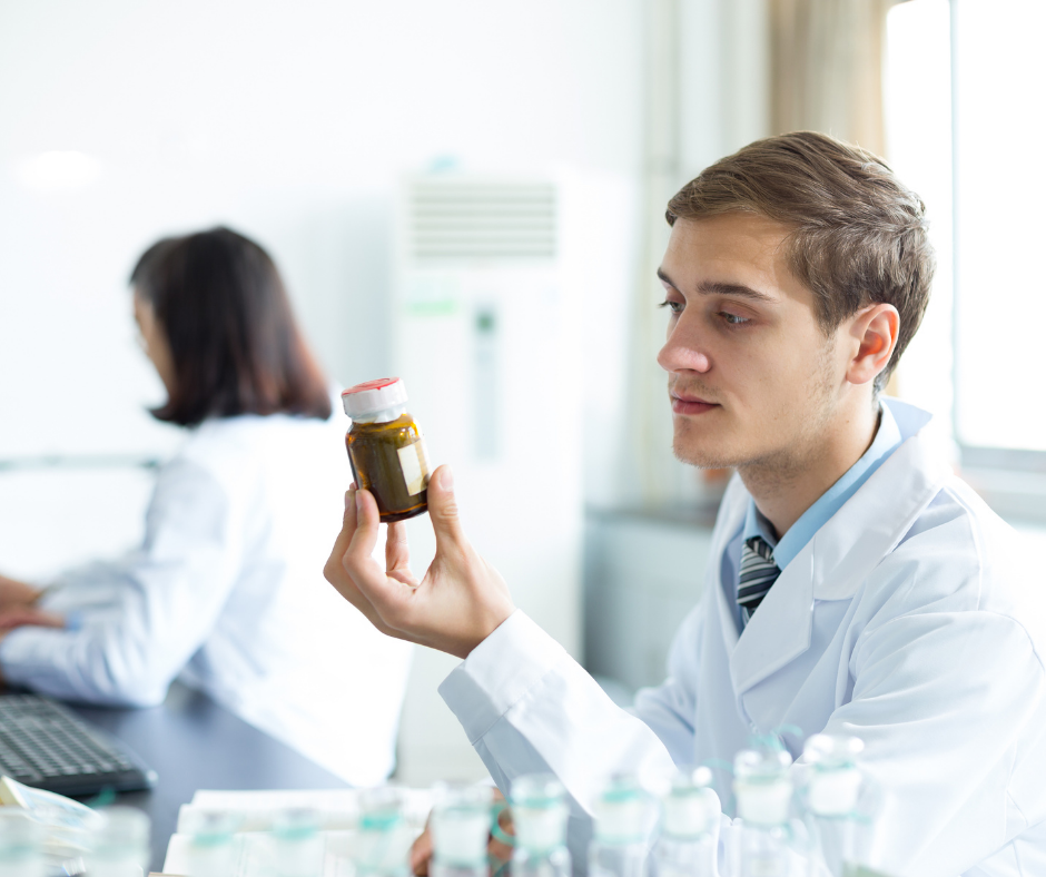 A person in a white lab coat examines a small brown bottle in a laboratory, with a colleague working in the background.