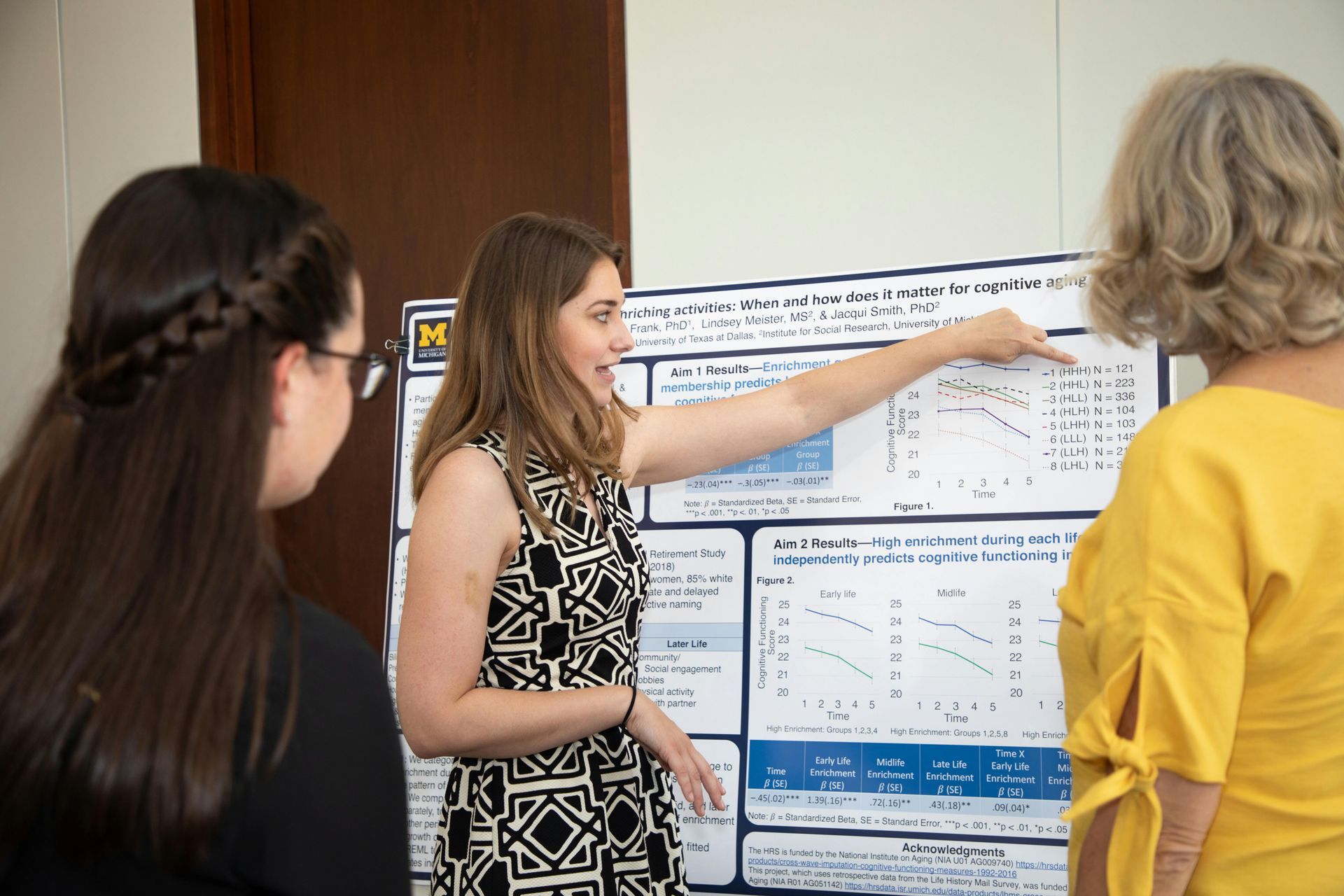 Three individuals stand around a research poster, one person gesturing to a graph as they present their findings.