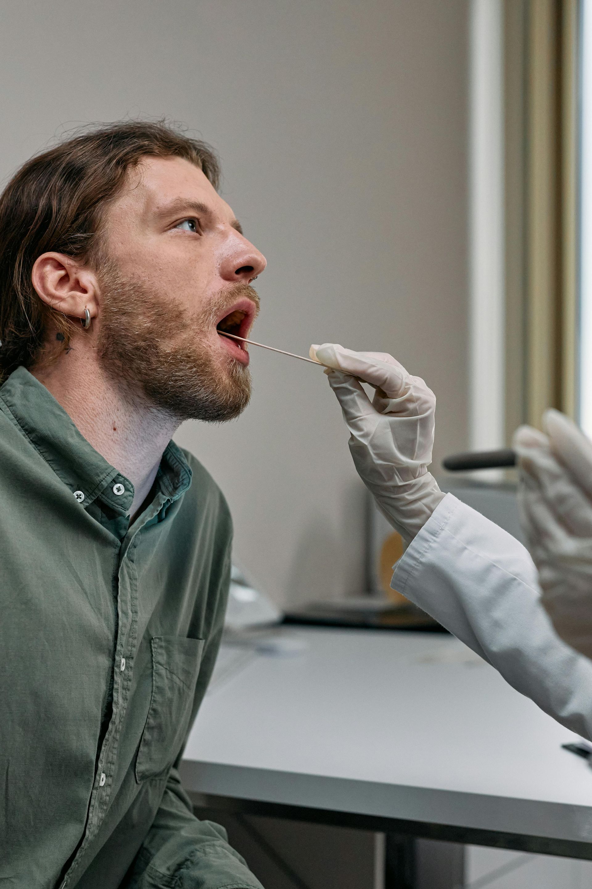 A healthcare worker in white gloves uses a swab to collect a sample from a person's mouth in a clinical setting.