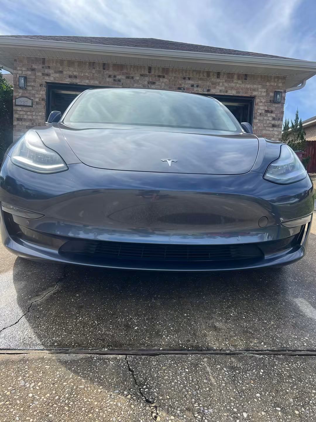Front view of a dark gray Tesla parked on a wet driveway in front of a stone house