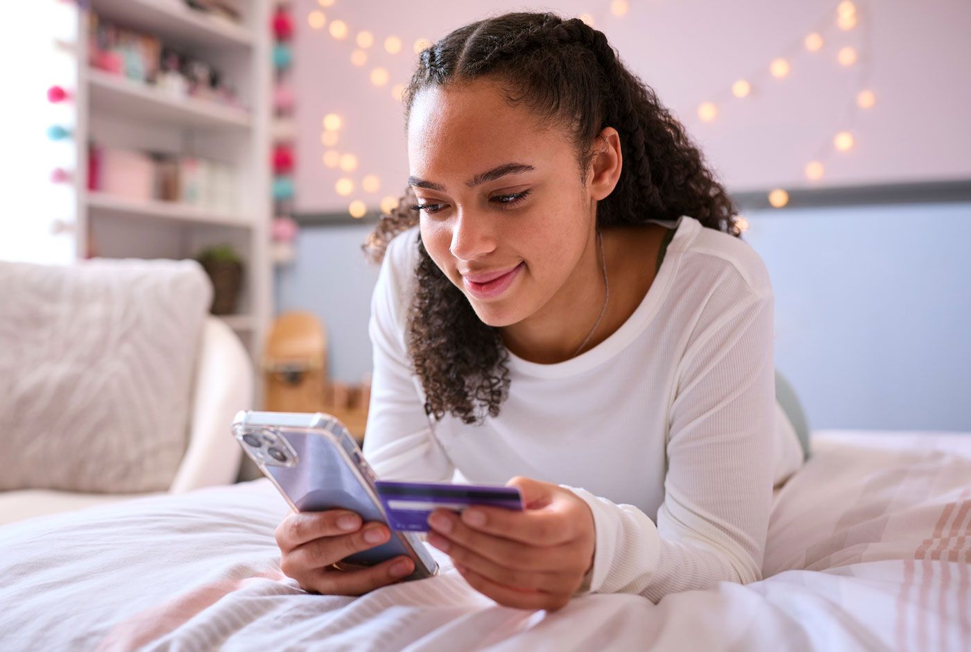 A person using a smartphone and credit card, smiling, lying on a bed in a room.
