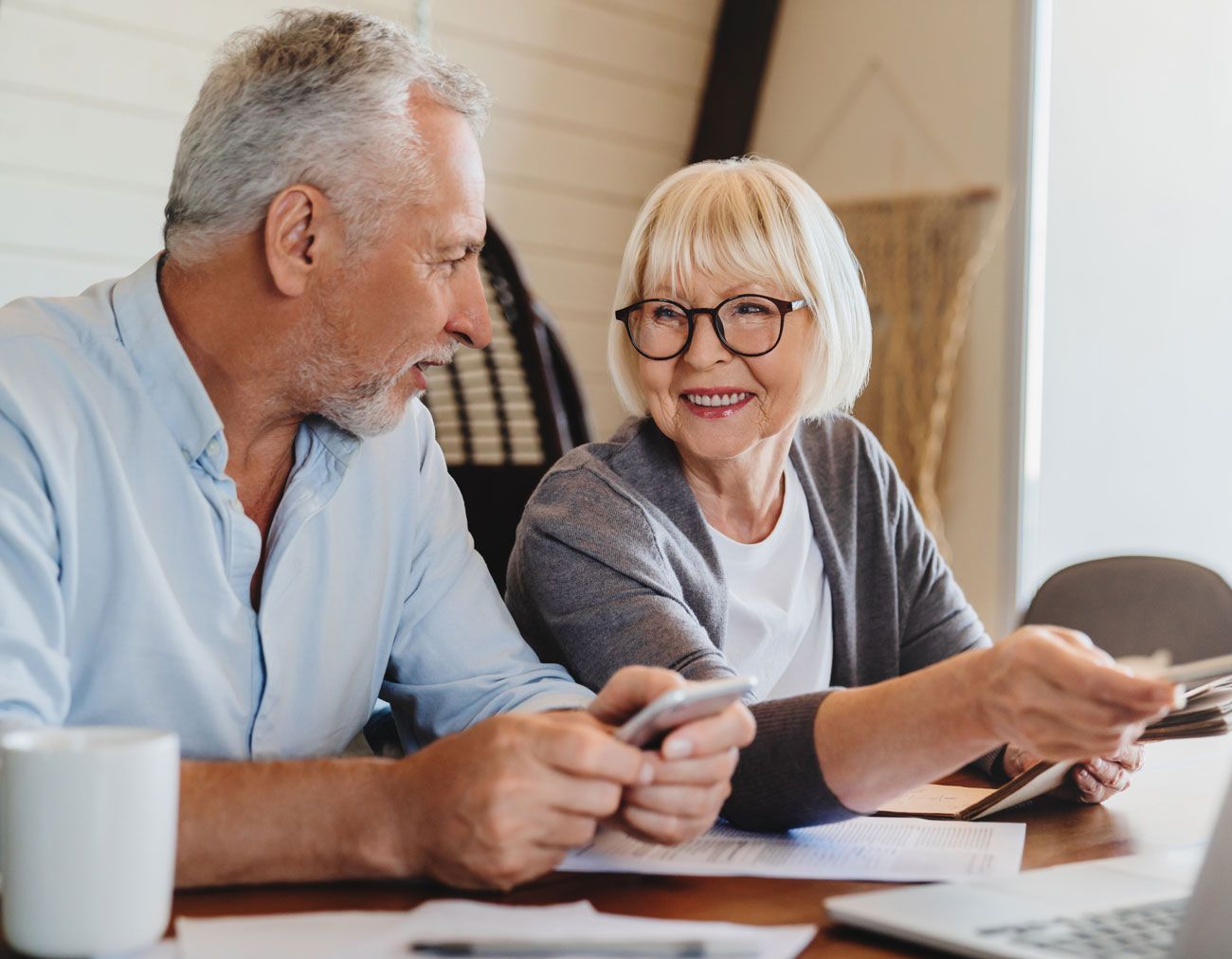 Smiling older couple reviewing papers and using a phone and laptop at a table.
