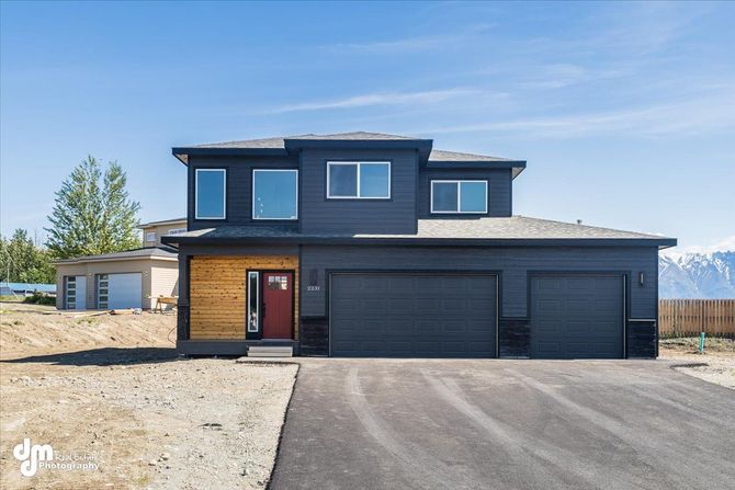 Modern two-story house with dark siding, red front door, and three-car garage on a gravel lot