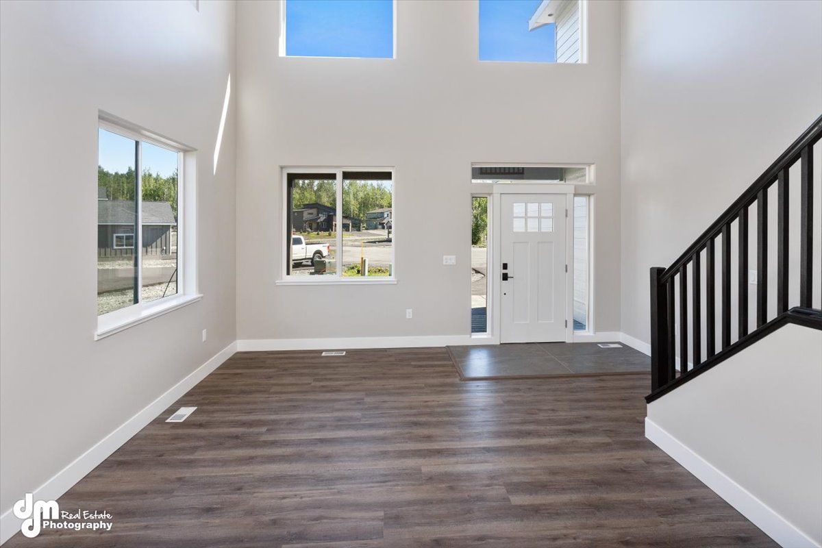 Bright empty living room with wood floors, white walls, tall windows, and a black staircase railing