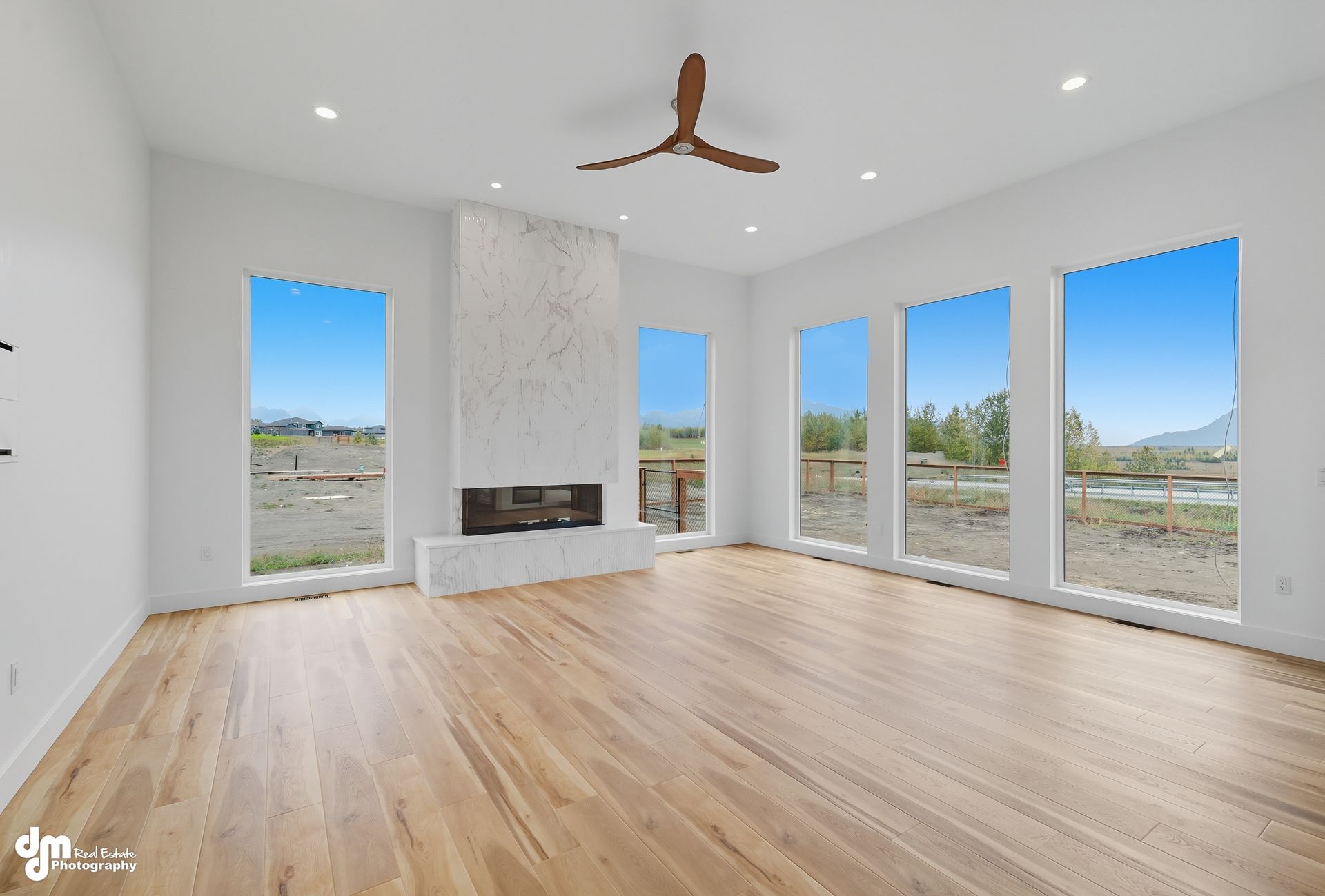 Bright open living room with wood floors, white walls, fireplace, ceiling fan, and large windows overlooking a landscape