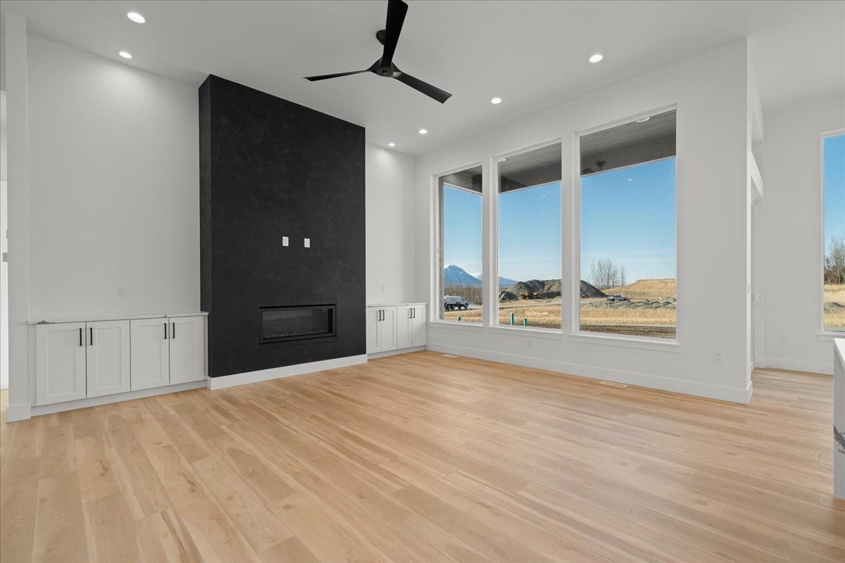 Empty modern living room with light wood floors, black fireplace wall, ceiling fan, and large windows.