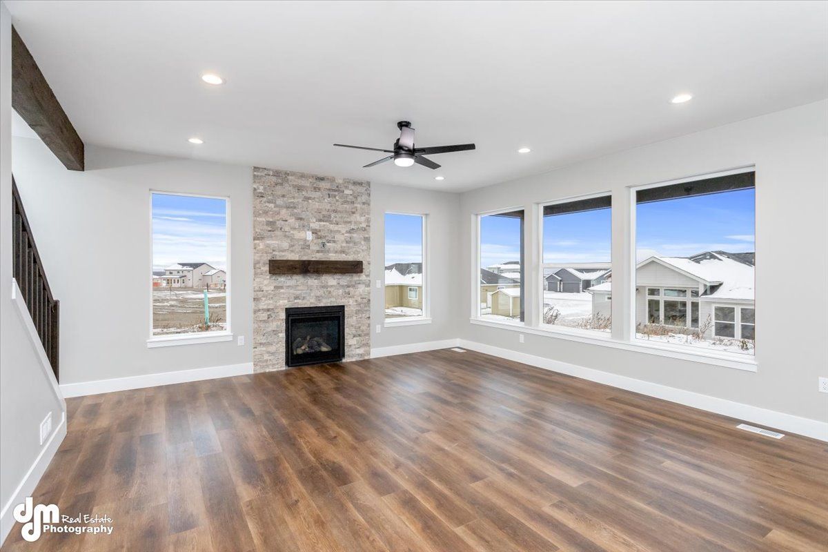 Bright empty living room with wood floors, stone fireplace, ceiling fan, and large windows.