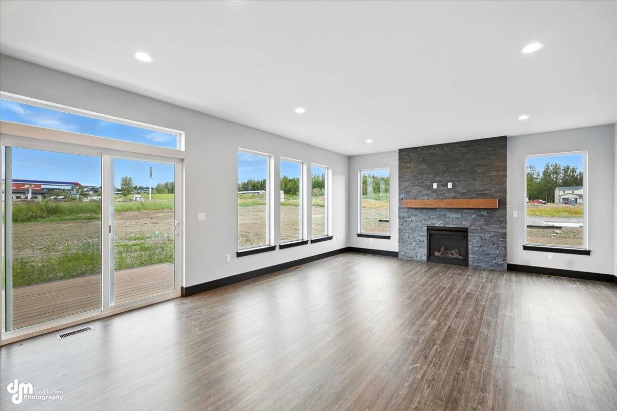 Empty modern living room with large windows, dark wood floors, and a stone fireplace.