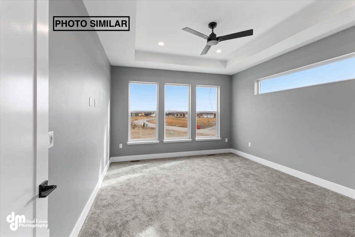 Empty gray bedroom with carpet, ceiling fan, and large windows overlooking a neighborhood.