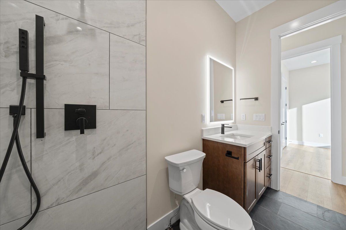 Modern bathroom with walk-in shower, black fixtures, vanity, and toilet leading to a bright hallway