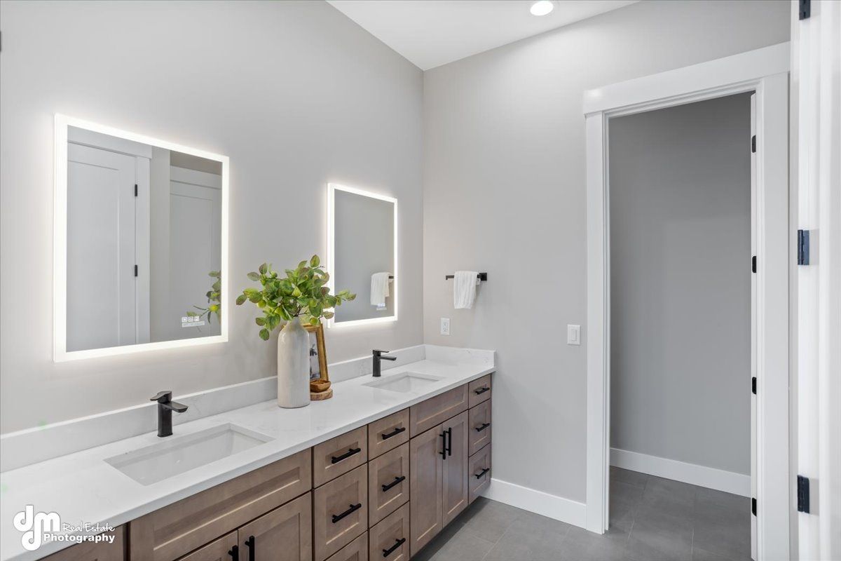 Modern bathroom vanity with double sinks, backlit mirrors, gray walls, and open doorway.