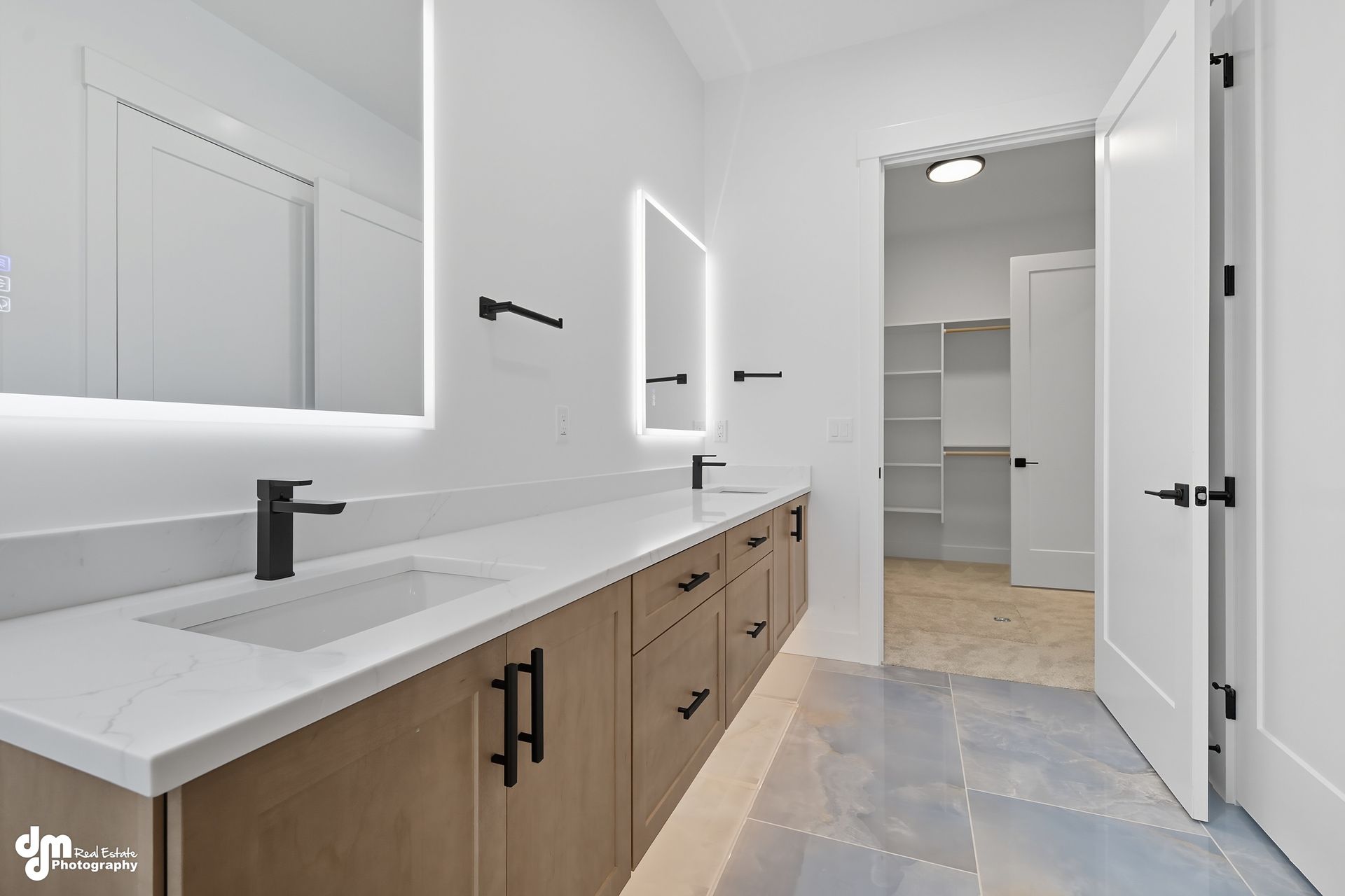 Bright modern bathroom with double vanity, black fixtures, and a walk-in closet beyond open doors.