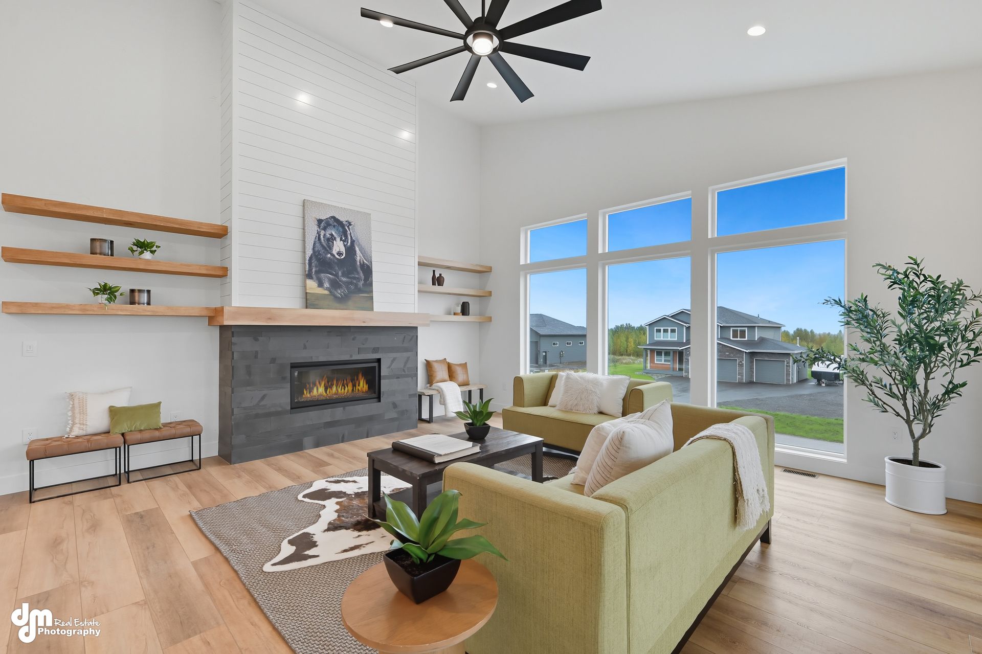 Bright living room with stone fireplace, light green sofas, wooden floors, and large windows overlooking a neighborhood.