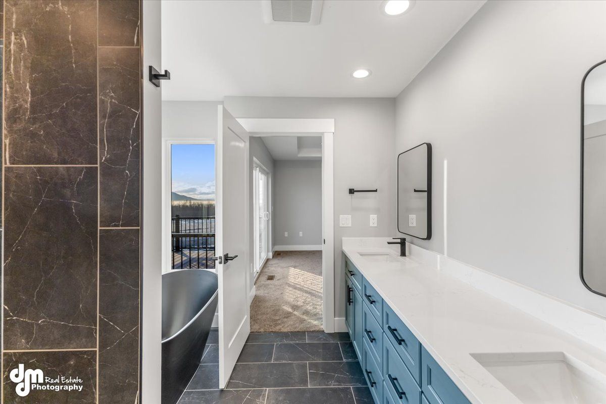 Long bathroom with blue vanity, white countertop, dark tile shower, and hallway view to a window.