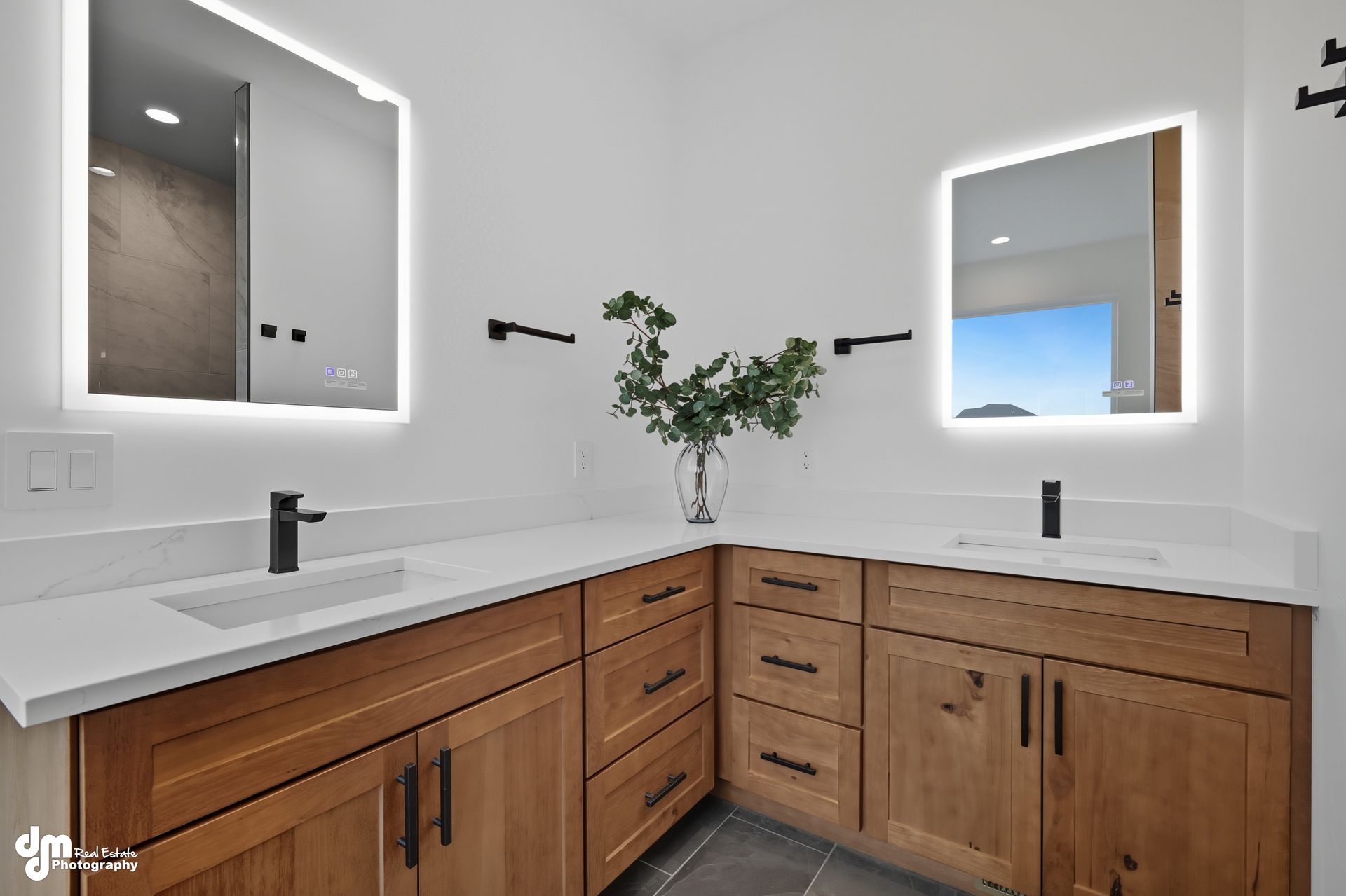 Modern bathroom vanity with wood cabinets, white countertop, black fixtures, and lit mirrors